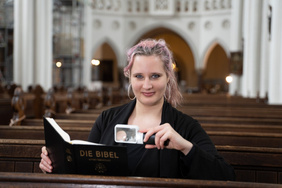 Eine Frau Mitte 20 sitzt in einer Kirchenbank und lächelt in die Kamera, in einer Hand eine Leuchtlupe, in der anderen eine Bibel. Sie hat langes, dunkelblondes Haar mit rötlichen Strähnchen und trägt große Ohrringe