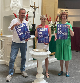 Kreispfarrer Torsten Maes, Pfarrerin Susanne Duwe und Pfarrerin Gesa Schaer-Pinne präsentieren das Plakat zur Taufe-to-go am 3.9.2022 in der Lambertikirche