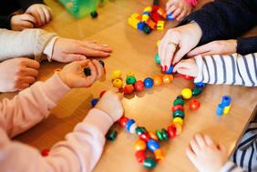 Kita - Kinder beim Spielen. An einem Tisch sitzen einige Kinder, eine Erzieherin und spielen mit einer Kette mit bunten Kugeln und anderen Formen, die auf einer Schnur aufgereiht sind.