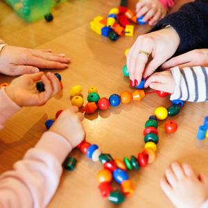 Kita - Kinder beim Spielen. An einem Tisch sitzen einige Kinder, eine Erzieherin und spielen mit einer Kette mit bunten Kugeln und anderen Formen, die auf einer Schnur aufgereiht sind.